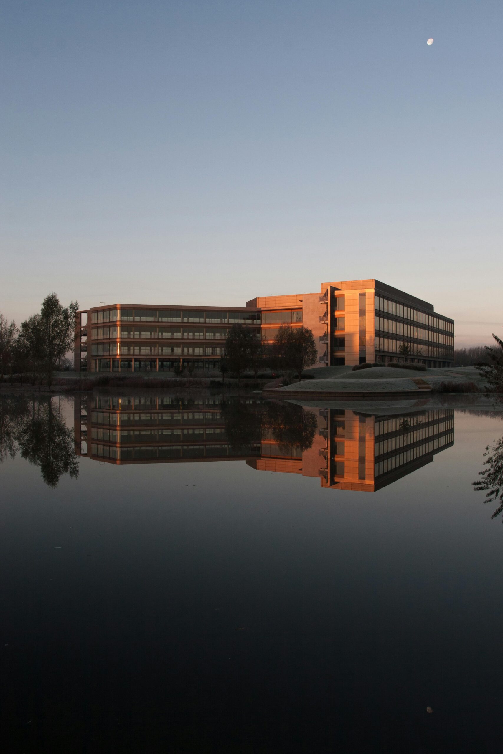 A modern office building reflected in a calm lake at sunset under a clear sky.
